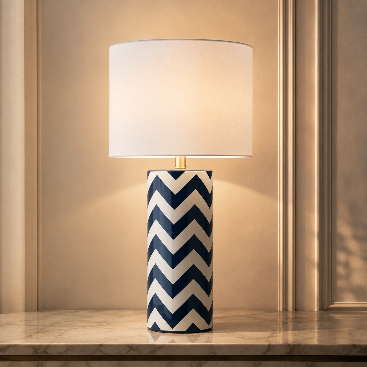A table lamp with a white drum shade and a cylindrical base featuring a navy and white chevron pattern, set on a marble surface against a warm beige paneled wall.