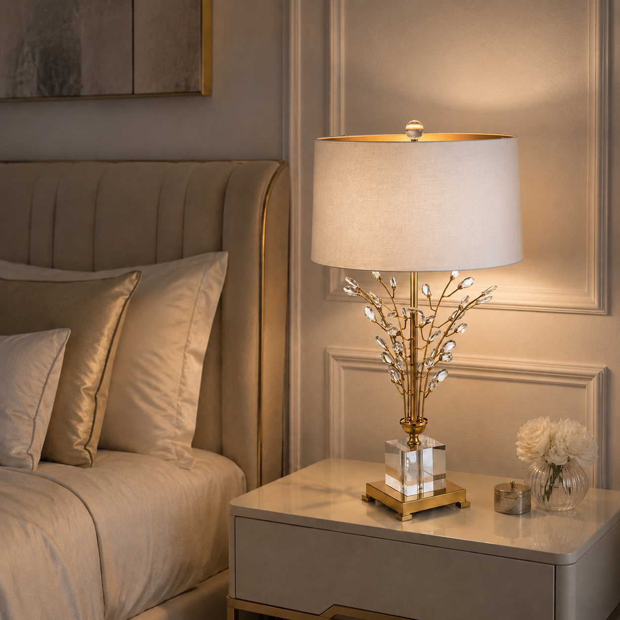 A luxury table lamp with a gold branch base and clear crystal accents, set on a marble nightstand next to a bed with beige bedding.