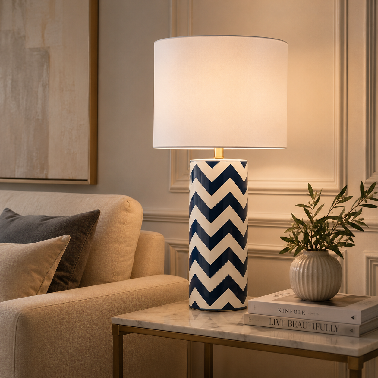 A table lamp with a white drum shade and a cylindrical base featuring a navy and white chevron pattern, styled on a marble-topped side table in a cozy living room setting.