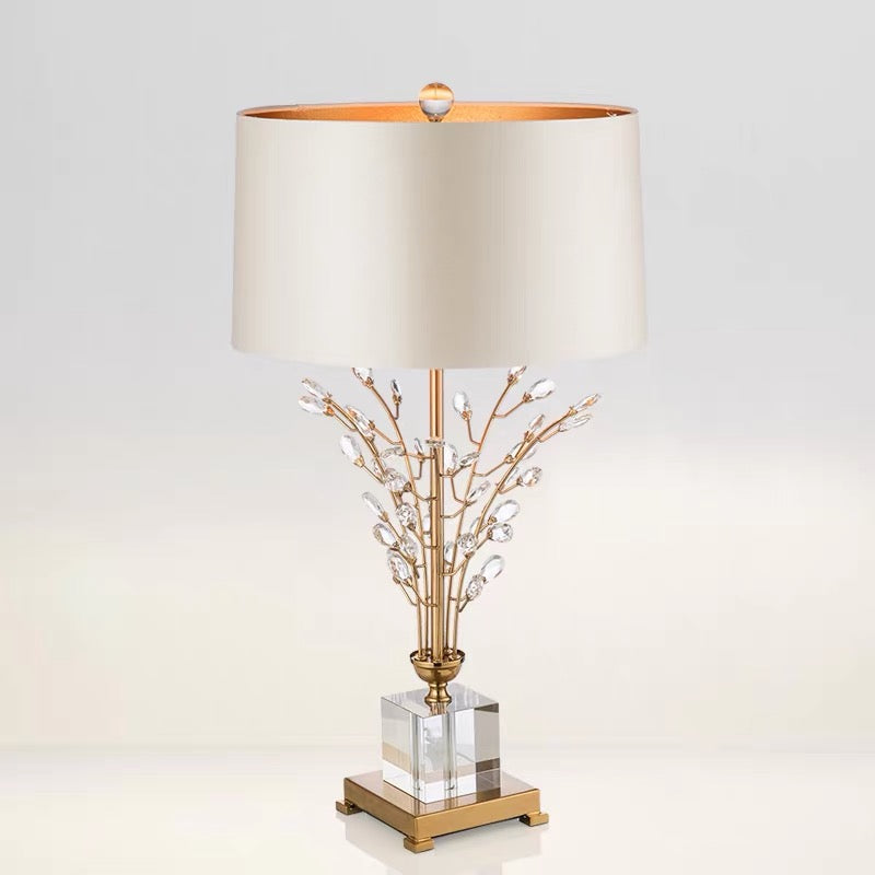 A table lamp with a large cream-colored fabric drum shade, a gold-finished metal frame with clear crystal branches, and a clear glass block base on a light gray background.