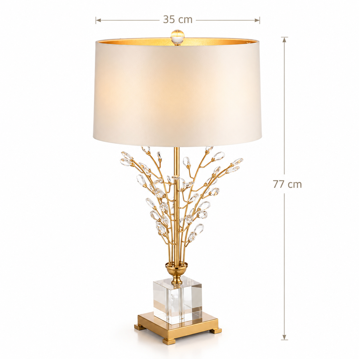 A table lamp with a warm white drum shade, a gold branch-like metal base adorned with clear crystal prisms, and a clear square glass block on a gold footed base. Measurement annotations show the shade width as 35 cm and the full lamp height as 77 cm.